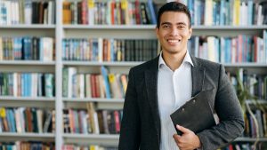 Young male standing in library in a suit with binder in hand