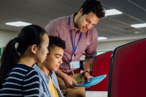 Students are working on computers with textbooks. A male teacher is standing with them, helping them with their work.