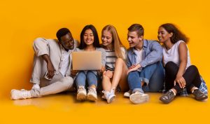 Carefree international teenagers sitting on floor and using laptop, yellow studio background, panorama