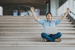 Asian male student celebrates sitting on steps with laptop