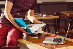 Body of teenager holding a book, sitting on a table with a laptop open.